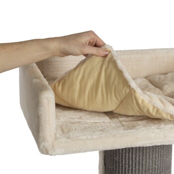 Eine Hand hebt ein abnehmbares, weiches, beiges Kissen von einem plüschigen Haustierbett mit einem cremefarbenen Äußeren, das sein gemütliches Design zeigt.