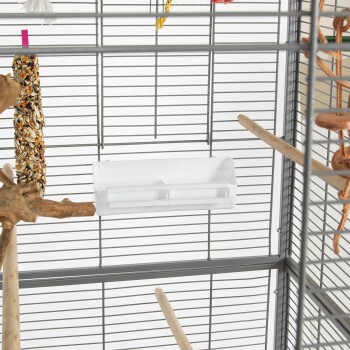 Das Innere eines Vogelkäfigs mit einer weißen Futterschale, Holzstangen und einem bunten Vogelsnack, der an der Seite hängt.