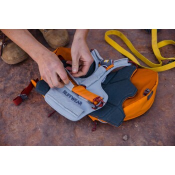 Eine Person passt ein leuchtend orangefarbenes Hundegeschirr mit grauer Polsterung und verstellbaren Riemen an, vor einem felsigen Untergrund.