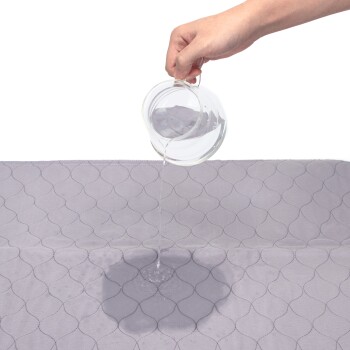 Eine Hand gießt Wasser aus einem Glasgefäß auf eine helllila Haustiermatte mit einem gesteppten Muster, um ihre wasserdichte Eigenschaft zu demonstrieren.