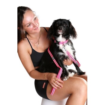 Eine Frau in einem schwarzen Tanktop und Shorts hält einen kleinen schwarz-weißen Hund mit einem rosa Geschirr und einer Leine vor einem weißen Hintergrund.