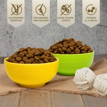 Zwei Schalen mit braunem Futter (gelb und grün) mit weißen Seilspielzeugen. Text: "Glutenfreie Rezeptur," "Für Gelenke," "Ohne Zucker," "Vitamin A, E, D₃."