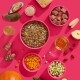 Ein lebendiger pinker Hintergrund zeigt eine zentrale Schüssel mit Tierfutter, umgeben von kleineren Schalen mit gewürfeltem Gemüse, Obst und einem Glas Öl.
