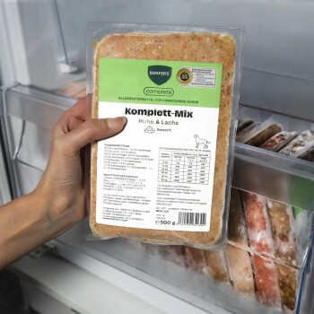 Hand hält verpacktes tiefgefrorenes Hundefutter mit der Aufschrift „Barfer's complete Komplett-Mix Huhn & Lachs 500 g“ im Gefrierschrank.