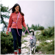 Eine Frau in einer roten Fleecejacke führt einen grau-weißen Husky an einer rosa Leine auf einem felsigen Weg, umgeben von Grün.