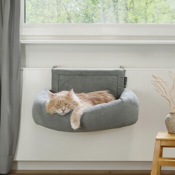 Eine flauschige orange Katze schläft friedlich in einem grauen hängenden Katzenbett, das am Fensterbrett befestigt ist, mit Grünpflanzen, die draußen sichtbar sind.