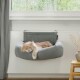 Eine flauschige orange Katze schläft friedlich in einem grauen hängenden Katzenbett, das am Fensterbrett befestigt ist, mit Grünpflanzen, die draußen sichtbar sind.