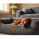 Rotbrauner Hund schläft in grauem Cord-Haustierbett mit Lederanhänger 'KNUFFELWUFF', im Hintergrund Sofa und Vase mit Tulpen.