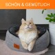 Eine flauschige orangefarbene Katze liegt in einem gemütlichen grauen Haustierbett und schaut mit leuchtend grünen Augen nach oben. Oben im Bild steht der Text 'SCHÖN & GEMÜTLICH.'