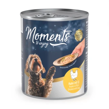 Une boîte de nourriture pour chiens étiquetée "Moments à savourer," avec un chien brun regardant une assiette de nourriture. Le texte indique "Huhn mit Ei" (Poulet avec œuf).
