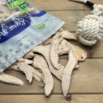 Getrocknete Hühnchen-Leckerlis verschüttet aus einem durchsichtigen Beutel auf Holz, neben einem weißen Tau-Spielzeug; Verpackungsaufschrift: 100% und Natürlich