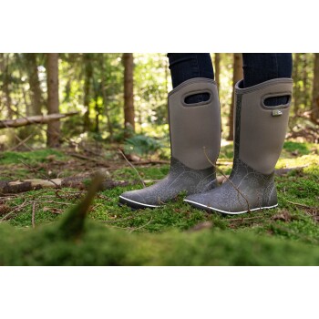 Ein Paar graue Gummistiefel mit Blumenmustern, getragen von einer Person, die auf moosigem Waldboden steht, umgeben von Bäumen.