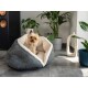 Ein kleiner Yorkshire Terrier sitzt bequem in einem gemütlichen grauen Haustierbett mit einem weichen weißen Innenraum, umgeben von Zimmerpflanzen und modernem Dekor.