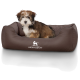 Ein kleiner, flauschiger Welpe sitzt bequem in einem braunen Hundebett mit einem gesteppten Innenraum, das das Logo "KNUFFELWUFF" trägt.