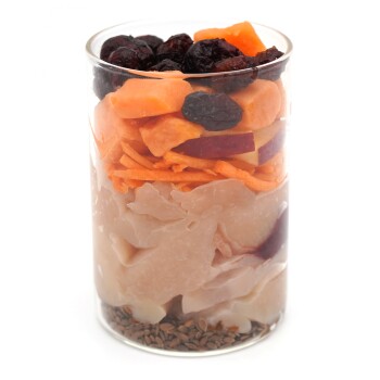 Ein klarer Glasbehälter mit Schichten von Haustierfutter: geriebene Süßkartoffel, getrocknete Cranberries, Fischscheiben und Leinsamen am Boden.