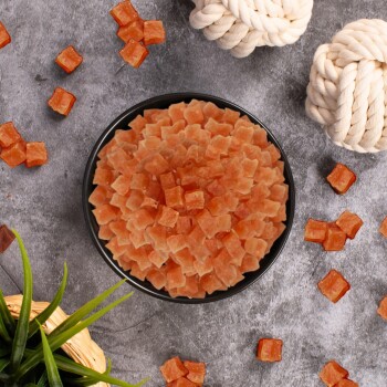 Eine schwarze Schale, gefüllt mit kleinen, orangefarbenen, würfelförmigen Hundeleckerlis, umgeben von verstreuten Leckerlis und zwei Seilspielzeugen auf einer grauen Oberfläche.