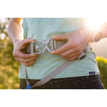 Eine Person, die eine graue Gürtelschnalle befestigt, während sie ein hellgrünes Hemd und gemusterte Shorts trägt, mit Sonnenlicht im Hintergrund.