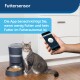 Eine graue getigerte Katze sitzt neben einem PetSafe-Futterautomaten auf Holzboden, während eine Hand ein Smartphone hält, das die "Futtersensor"-App für Futterwarnungen zeigt.