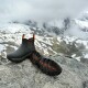 Schwarze und orangefarbene Hundestiefel mit robusten Sohlen auf einem Felsen, verschneite Berge im Hintergrund, beschriftet mit „Gaiter“ und „Trek“.