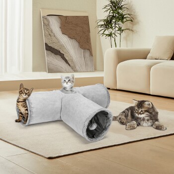 Zwei verspielte Katzen, eine getigerte und eine graue, erkunden einen weichen grauen Tunnel in einem gemütlichen Wohnzimmer mit einem beigen Sofa und einer Pflanze.