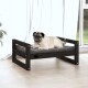 Ein Mops liegt bequem auf einem stilvollen schwarzen Holzhaustierbett mit einem grauen Kissen, in einem hellen Raum mit weißen Vorhängen und einer Pflanze.