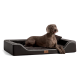 Ein brauner Hund liegt auf einem stilvollen schwarzen Haustierbett mit plüschigem Innenleben und einem Holzmarkenschild, vor einem dunklen Hintergrund.