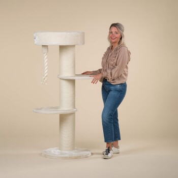 Eine Frau steht neben einem hohen, beigen Katzenbaum mit mehreren Ebenen, einem Kratzbaum und einem hängenden Seilspielzeug, vor einem neutralen Hintergrund.