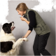 Eine Frau in einer grünen Weste interagiert mit einem großen, flauschigen schwarz-weißen Hund, hält seine Pfote und lächelt ihn an.