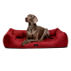Ein grauer Hund liegt auf einem plüschigen roten Haustierbett und schaut aufmerksam zur Seite. Das Bett hat ein schwarzes Etikett mit dem Markennamen.