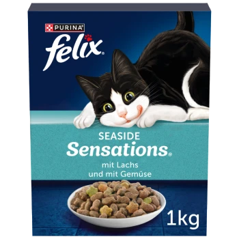 Un chat noir et blanc allongé à côté d'une boîte de nourriture pour chat Purina Felix Seaside Sensations, contenant des croquettes avec du poisson et des légumes, 1 kg.
