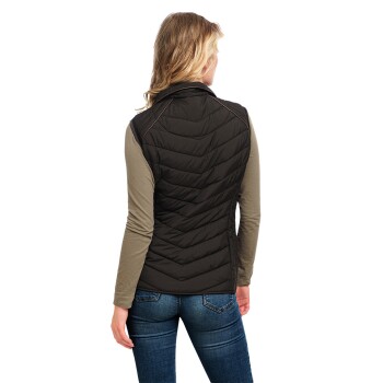 Eine Frau mit langen Haaren wird von hinten gesehen, trägt eine schwarze Steppweste über einem langärmeligen olivfarbenen Shirt und blauen Jeans.