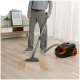 Eine Person in Grau saugt Krümel von einem Holzboden, mit einer Pflanze und einem gerahmten Bild im Hintergrund.