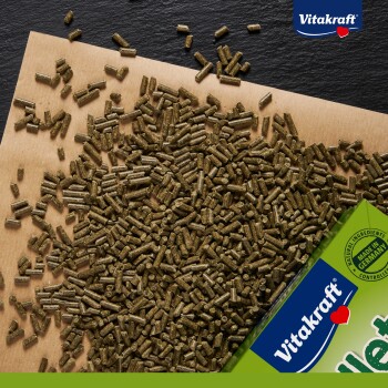Zbliżenie na zielone granulki karmy dla zwierząt na brązowej powierzchni, z rogiem opakowania Vitakraft pokazującym "Naturalne składniki" i "Wyprodukowano w Niemczech."