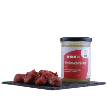 Ein Glas Rinderknochenbrühe mit der Aufschrift 'Fleischknochenbrühe' für Hunde und Katzen, begleitet von Stücken rohen Fleisches auf einer schwarzen Schieferoberfläche.