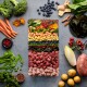 Eine bunte Anordnung von frischem Gemüse, Obst und Fleisch, darunter Brokkoli, Karotten, Heidelbeeren und verschiedene Fleischsorten, auf einem grauen Hintergrund.