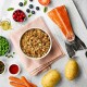 A bowl of pet food sits on a pink cloth, surrounded by fresh ingredients including salmon, peas, carrots, potatoes, and berries.
