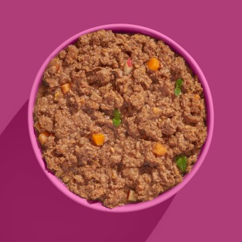 Eine rosa Schüssel gefüllt mit feuchtem Haustierfutter mit einer groben Textur, gemischt mit kleinen Stückchen Gemüse und Kräutern.