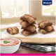 Ein Holzschneidebrett hält gestreifte Hundeleckerlis und eine Schüssel mit rosa Tiernahrung, die mit Schnittlauch garniert ist, mit dem Vitakraft-Logo.