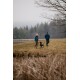 Eine Frau in einer grünen Jacke und ein Mann in einer blauen Jacke führen einen schwarzen Hund auf einem grasbewachsenen Weg in der Nähe eines Sees, umgeben von Bäumen.