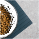 Eine schwarze Haustierschüssel, gefüllt mit trockenem Futter, steht auf einer dunkelblauen Matte, die mit "THE CATERY" geprägt ist, vor einer hellen strukturierten Oberfläche.