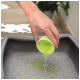 A hand pours green cat litter granules from a bright green cup into a gray litter box filled with gray litter.