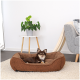 Ein kleiner brauner Hund, der sich in einem gemütlichen braunen Hundebett auf einem weichen Teppich entspannt, mit einer grünen Topfpflanze und einem hellgrauen Sofa im Hintergrund.