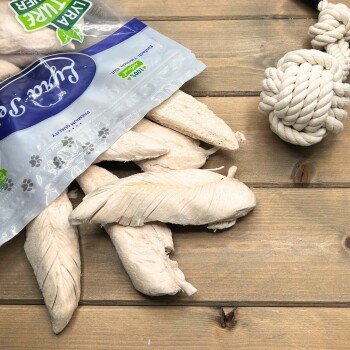 Weiße getrocknete Kausnacks, die aus einem Plastikbeutel auf einem Holztisch rieseln, neben einem geknoteten Seilspielzeug; '100%' sichtbar.