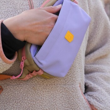 Eine Person in einer flauschigen beigen Jacke hält eine lavendelfarbene Leckerlitasche für Haustiere mit einem gelben Logo, in einer lässigen Outdoor-Umgebung.