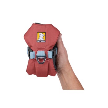 Eine Hand hält ein rotes Hundegeschirr mit einem blauen Riemen und einem metallenen D-Ring, mit einem gelben und schwarzen Ruffwear-Logo.