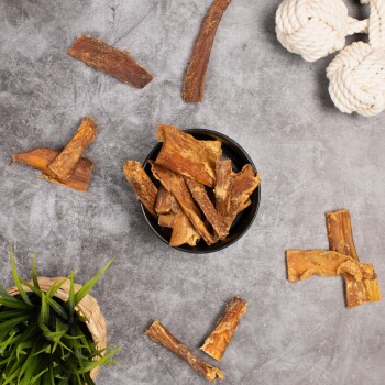 Eine schwarze Schüssel gefüllt mit Hundeleckerlis aus getrockneten Fleischstreifen, umgeben von verstreuten Stücken derselben Leckerlis und einer grünen Pflanze.