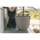 Eine graue Katze mit leuchtend gelben Augen schaut hinter einem gesprenkelten weißen Topf mit einer kleinen Pflanze hervor, vor einem Fensterblick.