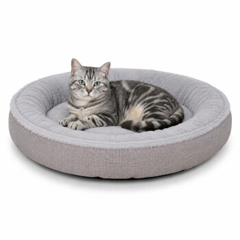 Grau getigerte Katze mit grünen Augen liegt in einem runden hellgrauen Plüsch-Tierbett mit erhöhtem Rand.