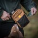 Person in grüner Jacke benutzt einen braun-schwarzen Hundeleckerlibeutel mit Metallflasche, zieht einen grünen Kotbeutel heraus; Hundeschnauze sichtbar.