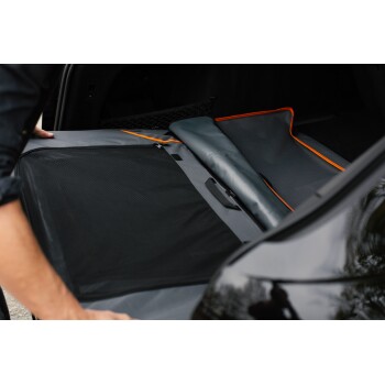 Eine Person entfaltet eine graue Autositzauflage für Haustiere mit orangefarbenem Rand im Kofferraum eines Fahrzeugs und bereitet sie zur Benutzung vor.